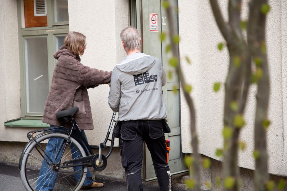 Två personer med en cykel vid en dörr i husets fasad. Den ena bär en jacka med Familjebostäders logotyp.